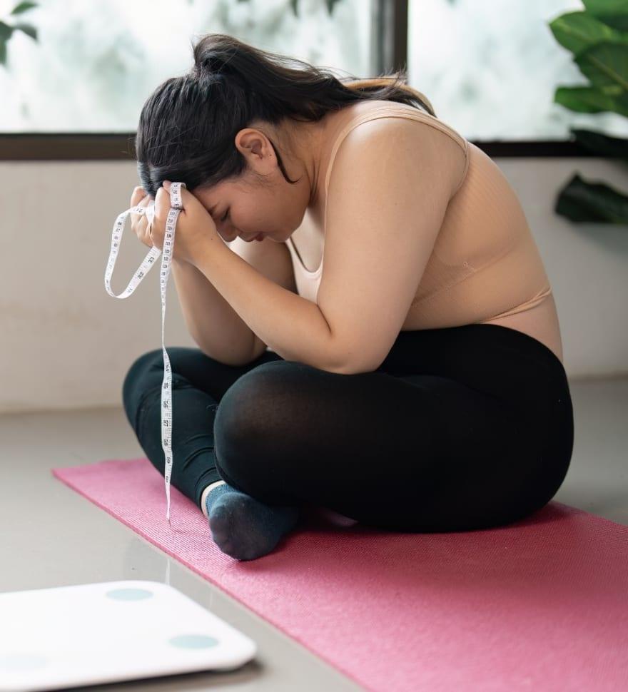 A woman sitting on a yoga mat holding a tape measure, frustrated