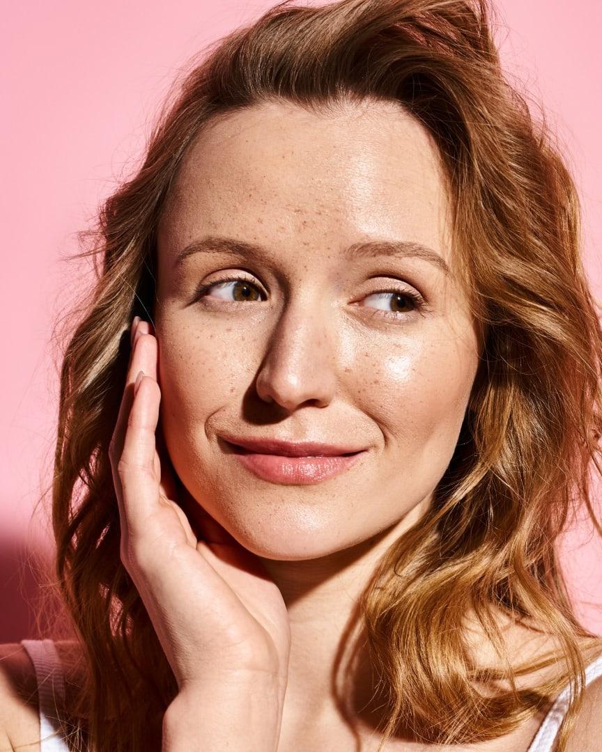 A woman with freckles and auburn hair touching her cheek, smiling